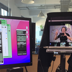 Behind-the-scenes view of a video recording setup, showing a person smiling and making a peace sign in the teleprompter screen.
